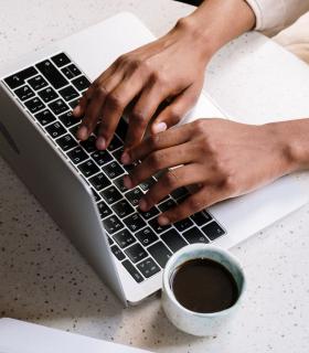 On voit des mains travaillant sur un clavier d'ordinateur portable accompagné d'un café. 